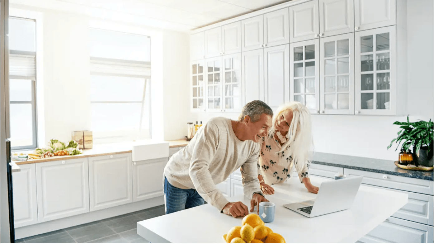 A couple is leaning over a laptop in a well lit kitchen.